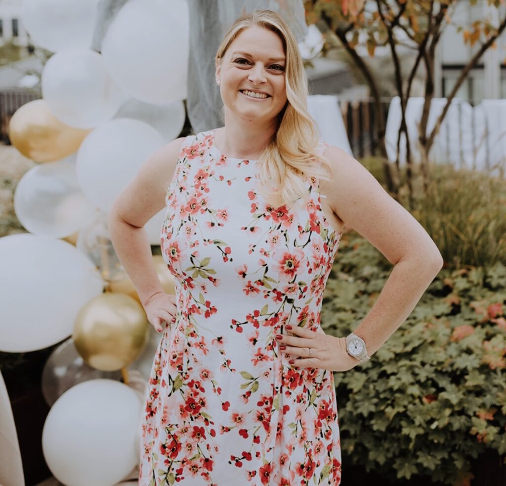 Anja Alberg lächelt in einem sommerlichen Blumen-Kleid vor gold-weißen Luftballons – eure Sängerin, Traurednerin und Hochzeitsplanerin.